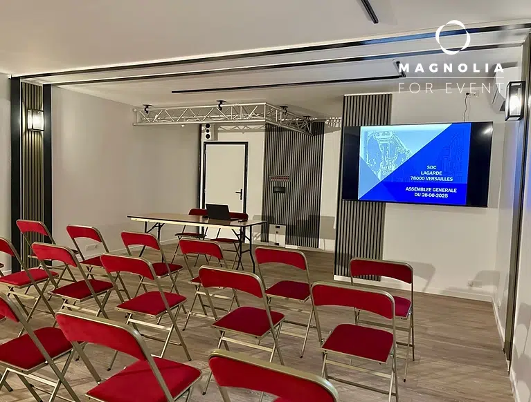 LE LAGARDE (78646) VERSAILLES : Une salle de conférence moderne, peu éclairée, avec des rangées de chaises pliantes rouges face à une table avec un ordinateur portable ; un écran affiche les détails de l'événement et "Assemblée Générale".
