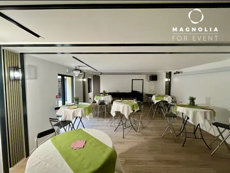 Une salle d'événement moderne présentant LE LAGARDE (78646) VERSAILLES, avec des tables rondes drapées de blanc et de vert, accompagnées de chaises et de petites plantes. Lumineux, sol en bois, et "Magnolia For Event" affiché sur le mur.