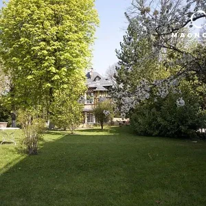 Le grand jardin herbeux du MANOIR DE LA CELLE SAINT CLOUD (78170) comprend de grands arbres, des buissons, une table et des chaises d'extérieur en métal blanc à gauche, et une maison avec des lucarnes sous un ciel bleu clair.