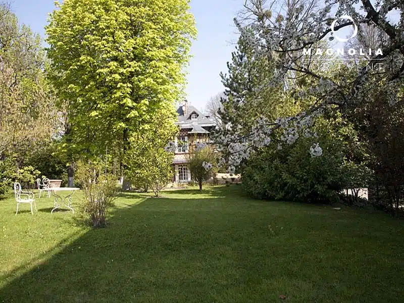 Le grand jardin herbeux du MANOIR DE LA CELLE SAINT CLOUD (78170) comprend de grands arbres, des buissons, une table et des chaises d'extérieur en métal blanc à gauche, et une maison avec des lucarnes sous un ciel bleu clair.