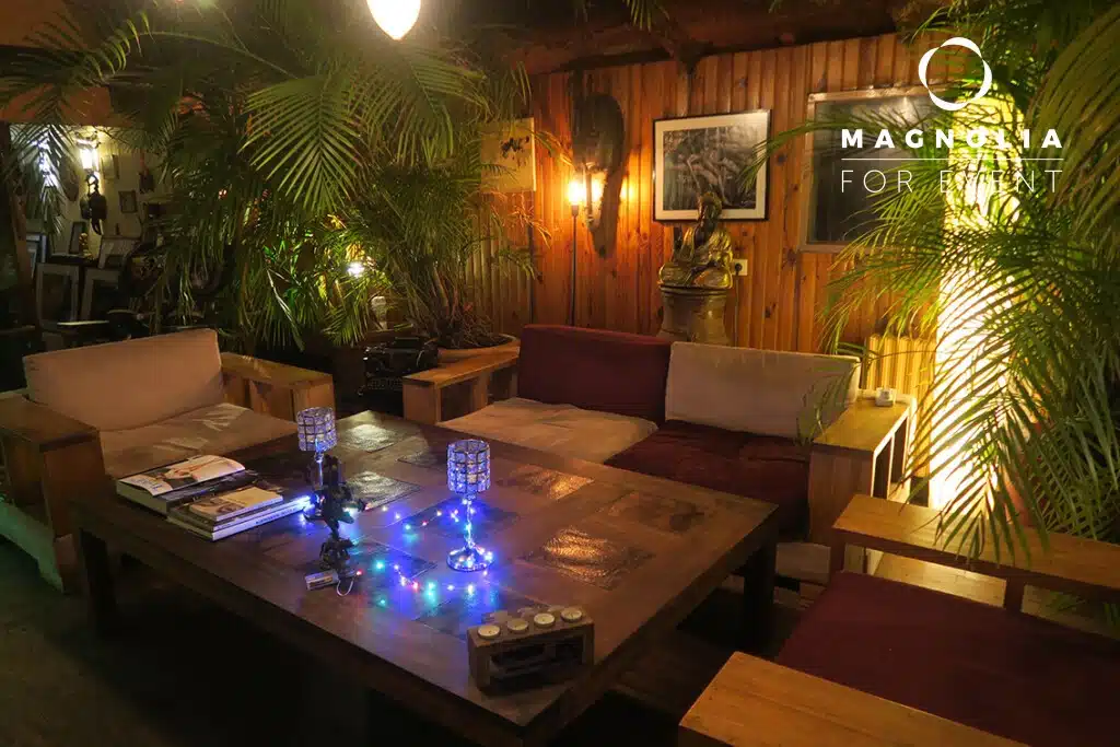 Un salon en bois confortable avec des canapés, une grande table basse, des plantes en pot, un éclairage doux et des œuvres d'art murales. Des livres, des bougies et des guirlandes lumineuses décorent la table. LA PÉNICHE SÉRÉNITÉ (92390) est inscrite dans le coin supérieur droit.