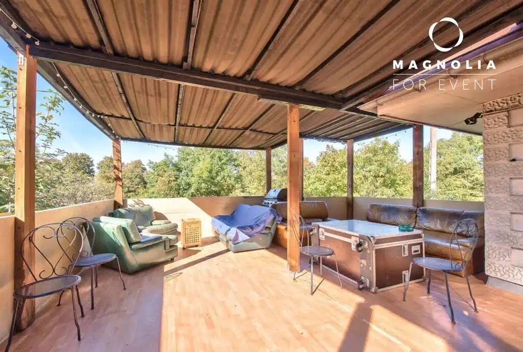 La terrasse extérieure couverte de LA DEMEURE DU BOIS DE VERRIERES (92140) est dotée d'un plancher en bois, de canapés, de fauteuils, de chaises en métal et d'une table. Elle est entourée d'arbres et baignée par la lumière du soleil.
