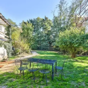 LA DEMEURE DU BOIS DE VERRIERES (92140) offre une cour ensoleillée avec une pelouse, une table en métal et six chaises, entourée d'arbres et de verdure. Un balcon et un mur couvert de lierre sont visibles sur le côté gauche de la maison.