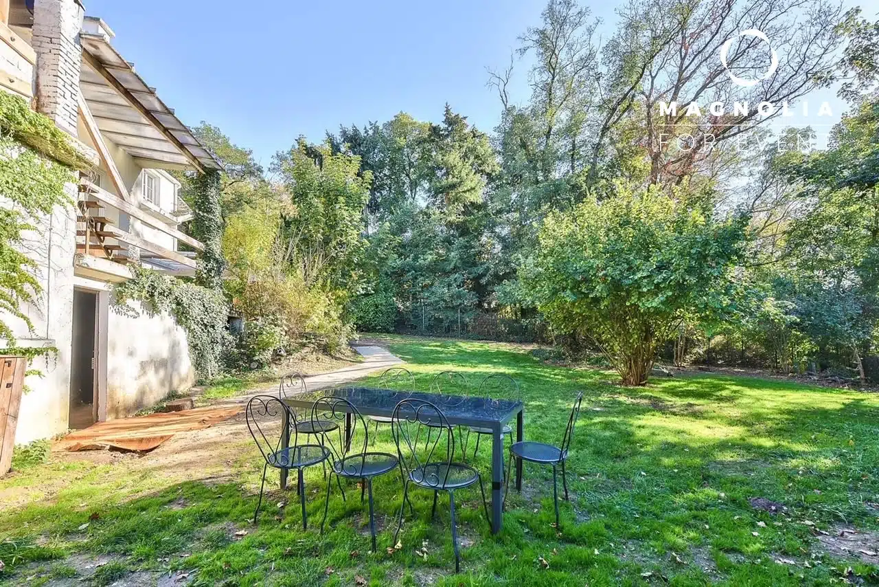 Sur une pelouse près d'une maison, la table d'extérieur LA DEMEURE DU BOIS DE VERRIERES (92140) en métal noir avec quatre chaises est placée au milieu d'arbres et de verdure sous un ciel bleu clair, avec le logo Magnolia dans le coin supérieur droit.