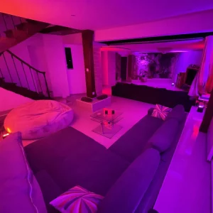Le salon confortable et futuriste du MANOIR DU 92 (92000) est doté d'un éclairage LED violet et rose, d'un canapé sectionnel avec des coussins rayés, d'une table basse en verre, d'un grand pouf et d'un escalier menant à l'étage.