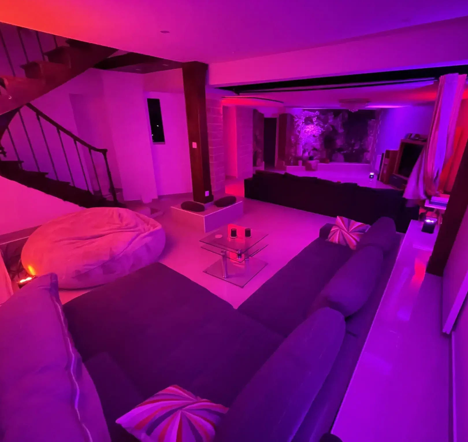 Le salon confortable et futuriste du MANOIR DU 92 (92000) est doté d'un éclairage LED violet et rose, d'un canapé sectionnel avec des coussins rayés, d'une table basse en verre, d'un grand pouf et d'un escalier menant à l'étage.