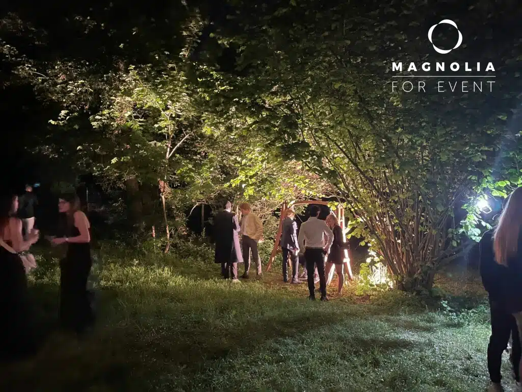 La nuit, les gens se rassemblent sous les arbres ornés de lumières brillantes à LA DEMEURE DU BOIS DE VERRIERES (92140), créant une scène festive en plein air. Certains invités se tiennent en petits groupes tandis que d'autres se promènent sur l'herbe. Le logo Magnolia For Event apparaît dans le coin supérieur droit.