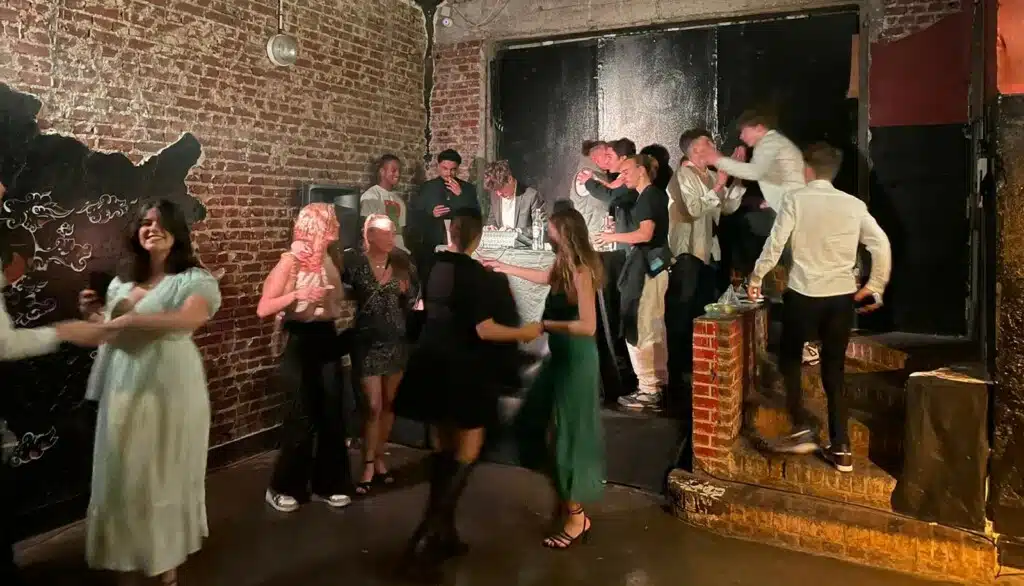 Des invités en tenue de fête dansent et se rencontrent dans LA DEMEURE DU BOIS DE VERRIERES (92140), faiblement éclairée, aux murs de briques apparentes et à l'ambiance festive. Certains se mêlent à la foule sur une plate-forme surélevée, tandis que d'autres profitent de la scène animée sur le sol.