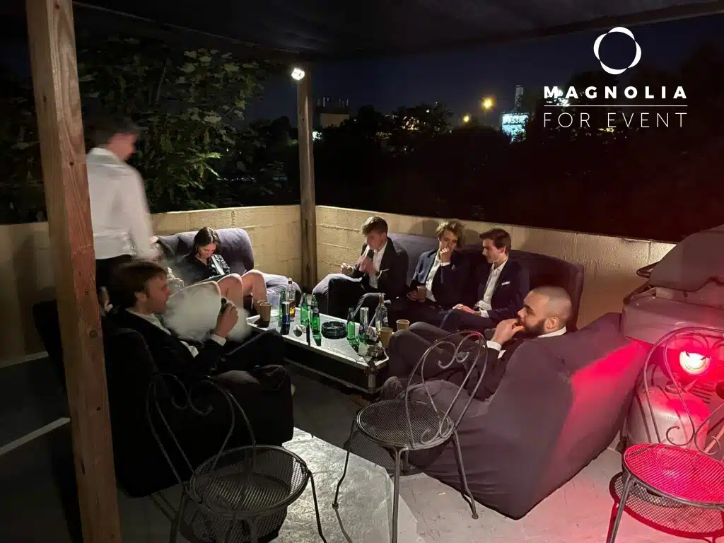 Un groupe en tenue de soirée se détend sur des canapés avec des boissons autour d'une table sur une terrasse extérieure le soir à LA DEMEURE DU BOIS DE VERRIERES (92140), avec une personne debout. Le logo de l'établissement est visible dans le coin supérieur droit.