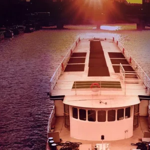 LE BOREAS (PARIS) navigue sur une rivière au coucher du soleil, le soleil orange bas sur l'horizon se reflétant dans l'eau. Le pont du LE BOREAS est visible au premier plan, des silhouettes de villes apparaissent à l'arrière-plan.