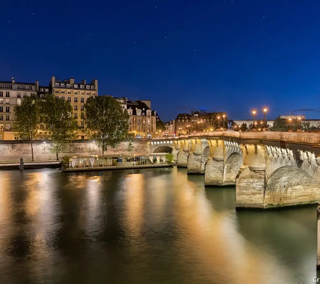 Vue nocturne de LA PÉNICHE LOCET (75013) à Paris, avec des bâtiments illuminés et des lampadaires se reflétant sur la Seine calme sous un ciel bleu profond.