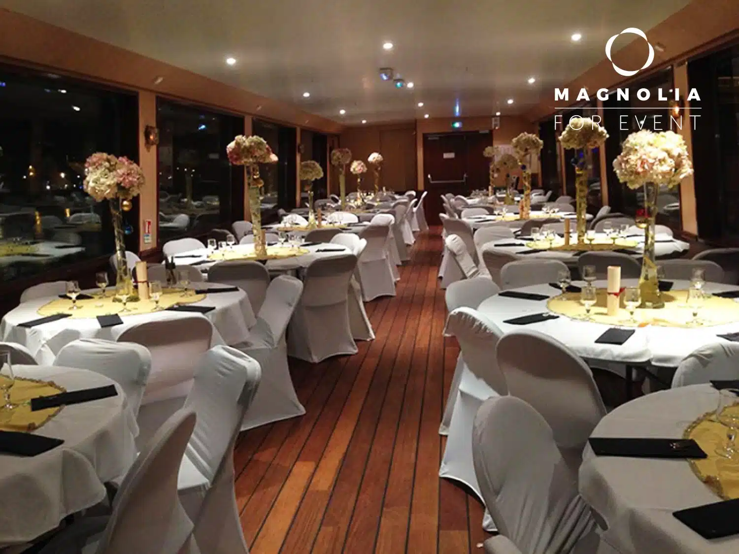 LA PÉNICHE LOCET (75013) dispose d'une salle de banquet élégante avec des tables rondes en lin blanc, des centres de table en fleurs dorées, des chaises blanches, un éclairage chaleureux, des planchers en bois et un logo Magnolia for Event dans le coin supérieur droit.