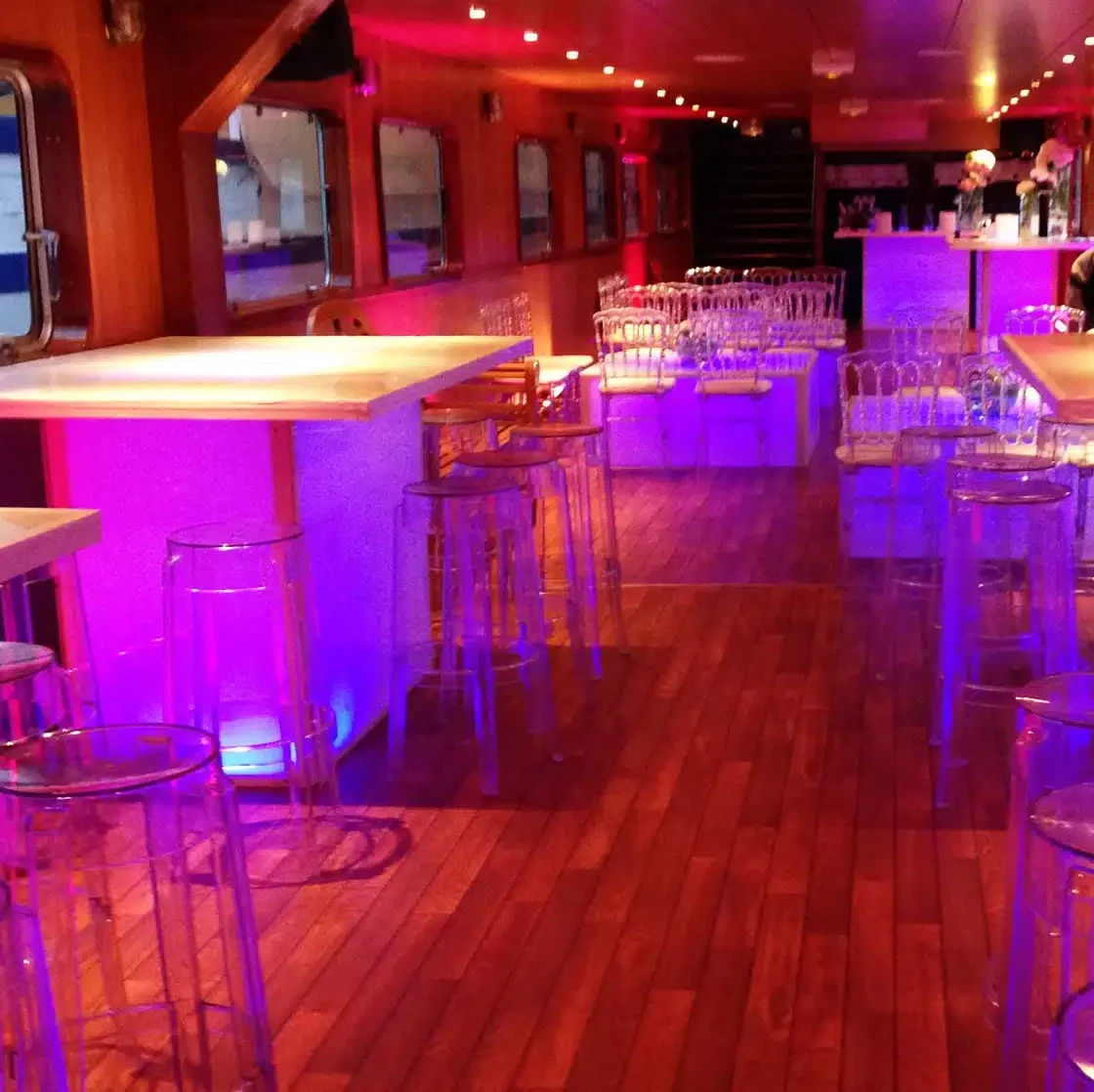 LA PÉNICHE LOCET (75013) est un bar élégant avec des planchers en bois, des tables hautes, des chaises transparentes, un éclairage rose et violet vibrant, un espace vide, un bar à l'arrière et des fenêtres sur un côté.