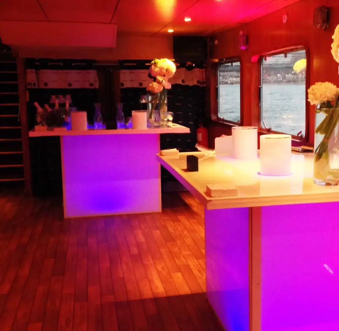 LA PÉNICHE LOCET (75013) propose un bar intérieur élégant avec des comptoirs éclairés de violet, des vases de fleurs blanches, des planchers en bois et de grandes fenêtres donnant sur l'eau ; un éclairage doux crée une atmosphère chaleureuse.