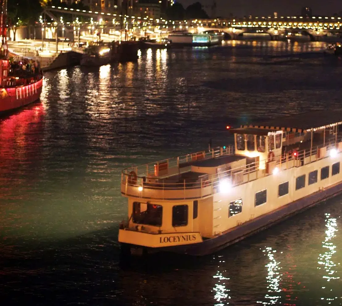 Un bateau fluvial blanc nommé LA PÉNICHE LOCET (75013) glisse le long d'un fleuve de nuit, illuminé par ses feux. L'eau reflète les lumières de la ville et les ponts lointains à l'arrière-plan.