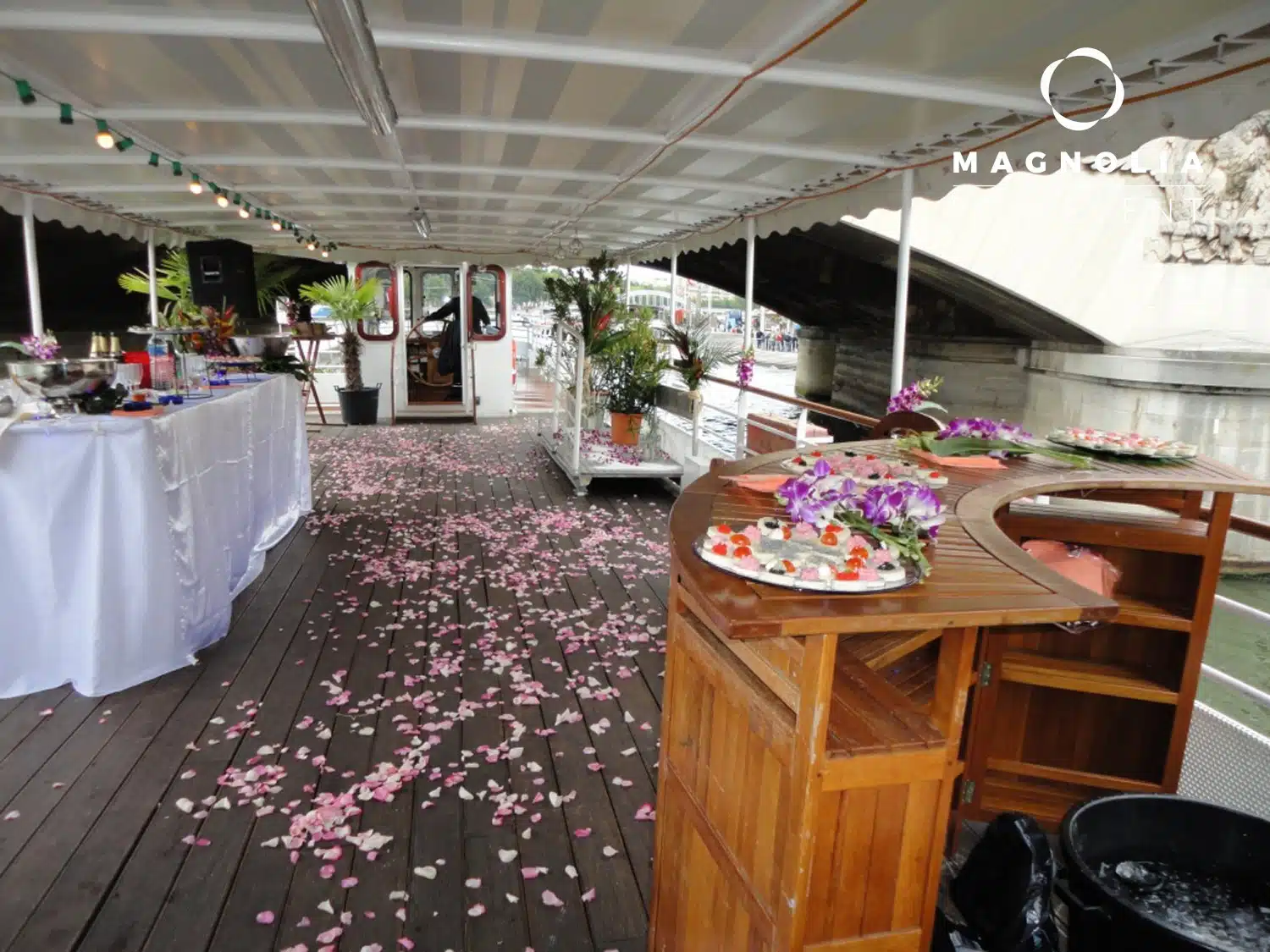 Une terrasse décorée de LA PÉNICHE LOCET (75013) présente des tables vêtues de blanc, des pétales de fleurs sur le sol en bois, des compositions florales et des plateaux de hors-d'œuvre ; "MAGNOLIA" apparaît en haut à droite.