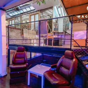 LE LOFT DE TITO (75011) est un espace événementiel moderne avec un éclairage violet, deux chaises en cuir marron autour d'une table blanche, un escalier en métal, un niveau supérieur vitré avec des chaises en bois, et le logo Magnolia For Event en vue.