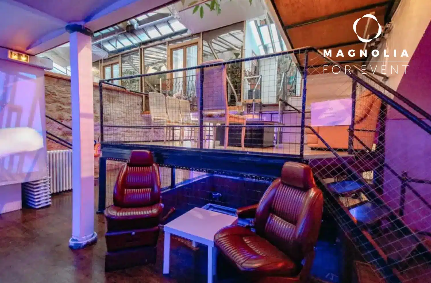 LE LOFT DE TITO (75011) est un espace événementiel moderne avec un éclairage violet, deux chaises en cuir marron autour d'une table blanche, un escalier en métal, un niveau supérieur vitré avec des chaises en bois, et le logo Magnolia For Event en vue.