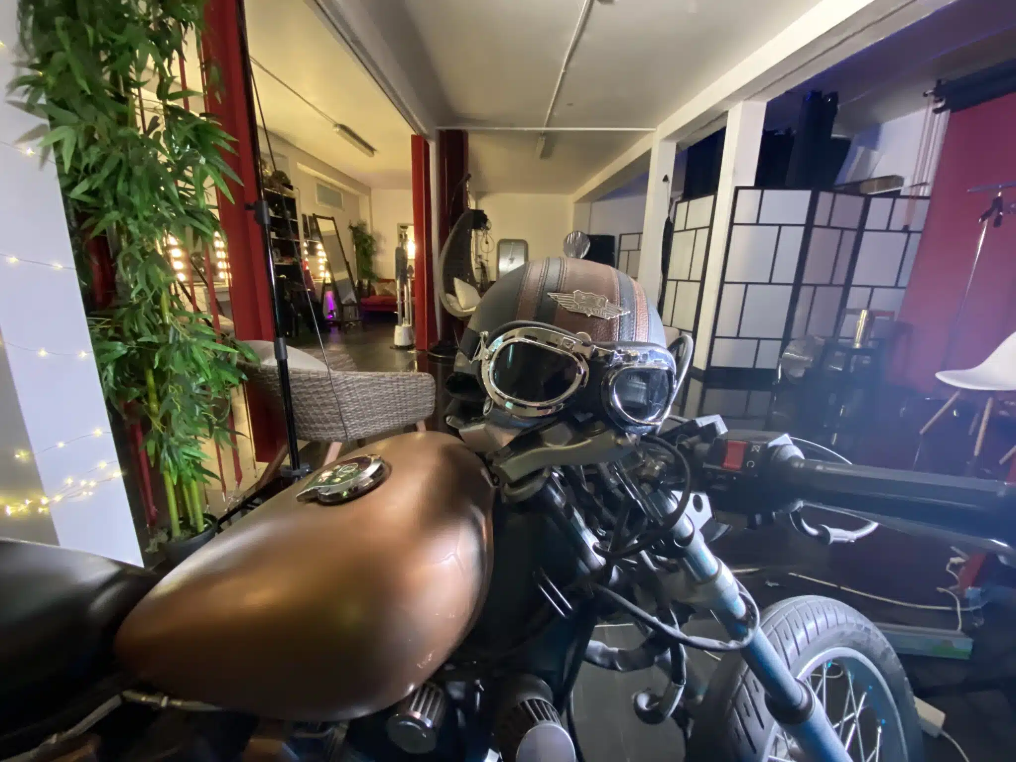 Gros plan sur une moto marron avec des lunettes d'aviateur sur le siège, à l'intérieur de LE LOFT DU 20ÈME (75020), avec en arrière-plan des plantes, des guirlandes lumineuses et des meubles modernes.
