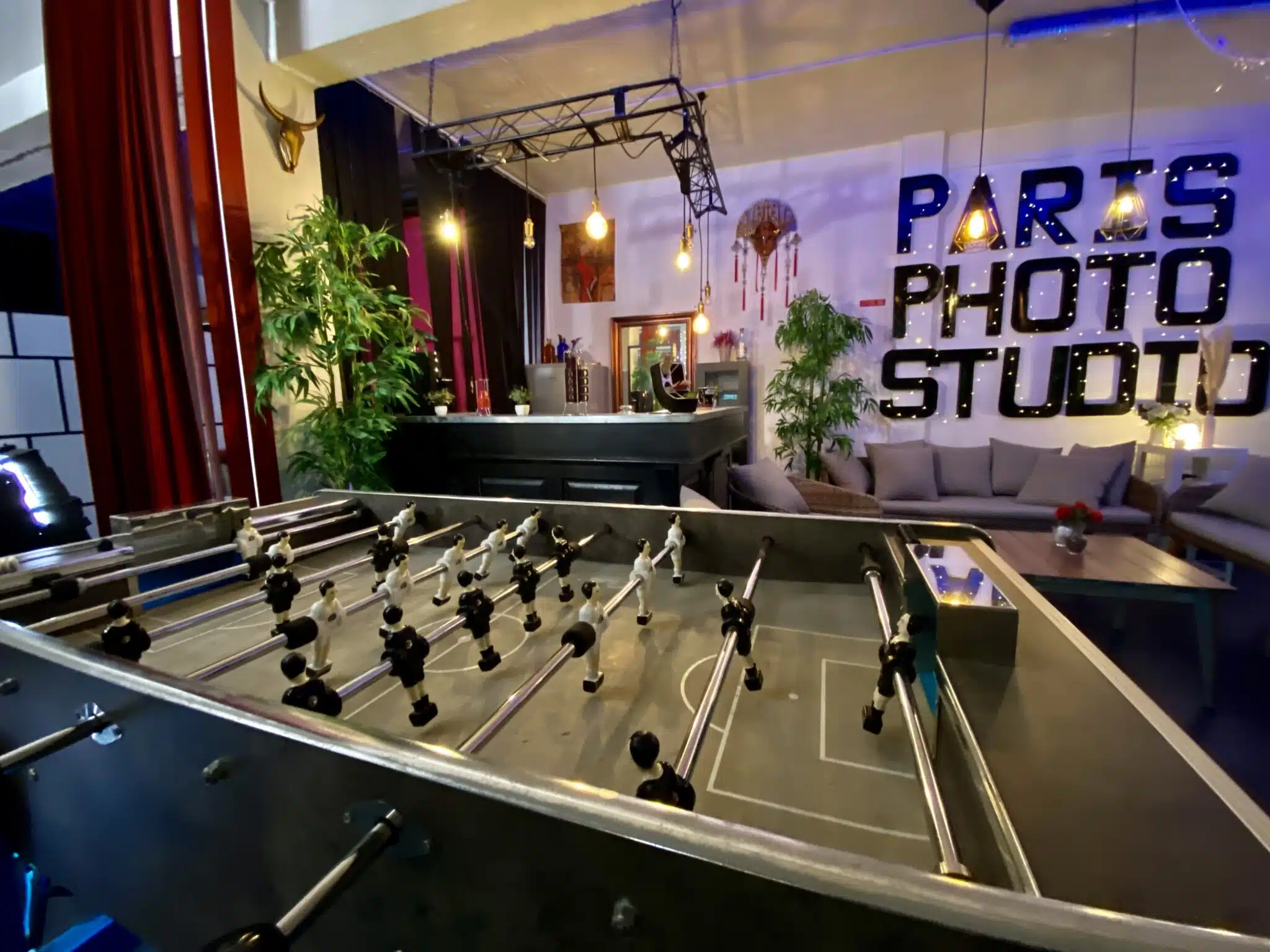 LE LOFT DU 20ÈME (75020) dispose d'un espace intérieur élégant avec un baby-foot, des plantes luxuriantes, un bar, des sièges confortables et une enseigne murale audacieuse "Paris Photo Studio". Un éclairage chaleureux crée une ambiance détendue et accueillante.