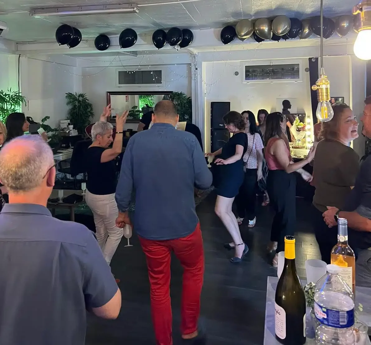 Un groupe animé danse et socialise au LOFT DU 20ÈME (75020), avec des ballons noirs au-dessus, des bouteilles de vin sur la table et une atmosphère festive à l'intérieur.
