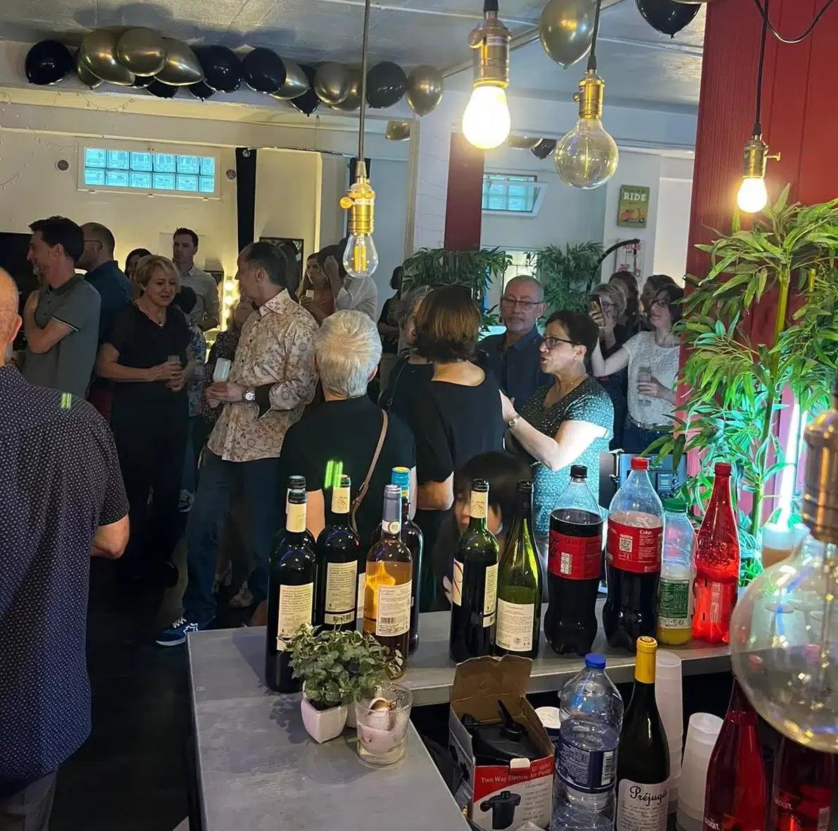Au LOFT DU 20ÈME (75020), les gens se rencontrent lors d'une fête à l'intérieur. Des bouteilles de vin, de soda et d'eau sont posées sur un comptoir, tandis que des lampes suspendues, des plantes et des ballons décorent l'espace animé.
