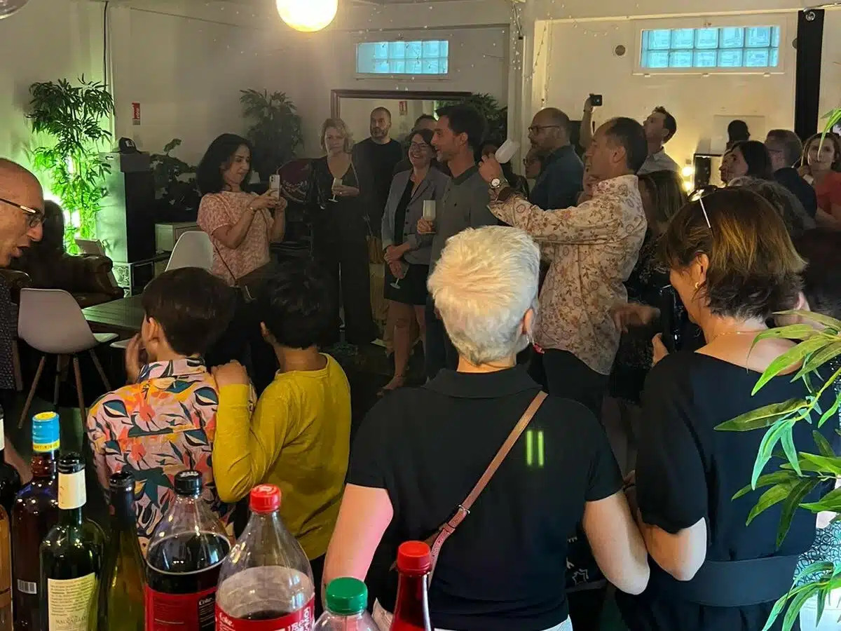 Un groupe se réunit à l'intérieur du LOFT DU 20ÈME (75020) pour un événement social. Certains discutent pendant que d'autres prennent des photos. Des boissons et des bouteilles sont disposées sur une table au premier plan, avec un éclairage chaud et des plantes en arrière-plan.