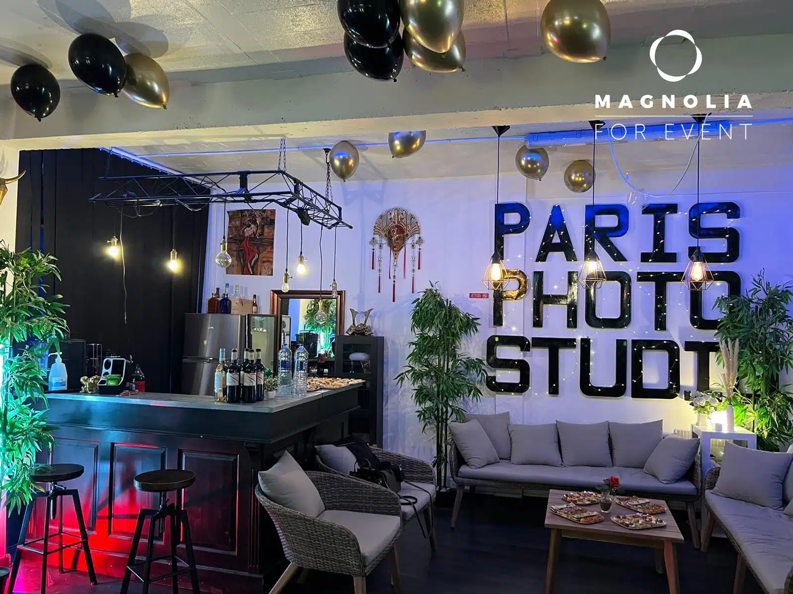 Un salon intérieur festif au LOFT DU 20ÈME (75020) avec un bar, un coin salon, des plantes, des ballons dorés et noirs, et de grandes lettres noires PARIS PHOTO STUDIO sur le mur. Un logo Magnolia For Event apparaît dans le coin supérieur droit.