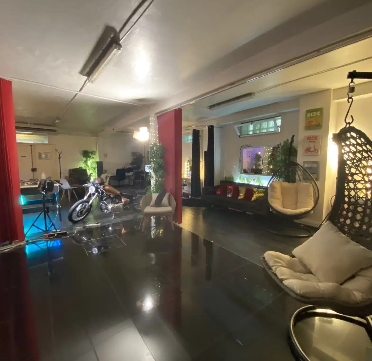 LE LOFT DU 20ÈME (75020) présente une pièce moderne et spacieuse avec un sol noir brillant, un fauteuil œuf suspendu, un canapé, des plantes, des lumières colorées, une moto et des rideaux rouges. Un décor élégant et un éclairage d'ambiance doux créent une atmosphère chaleureuse.