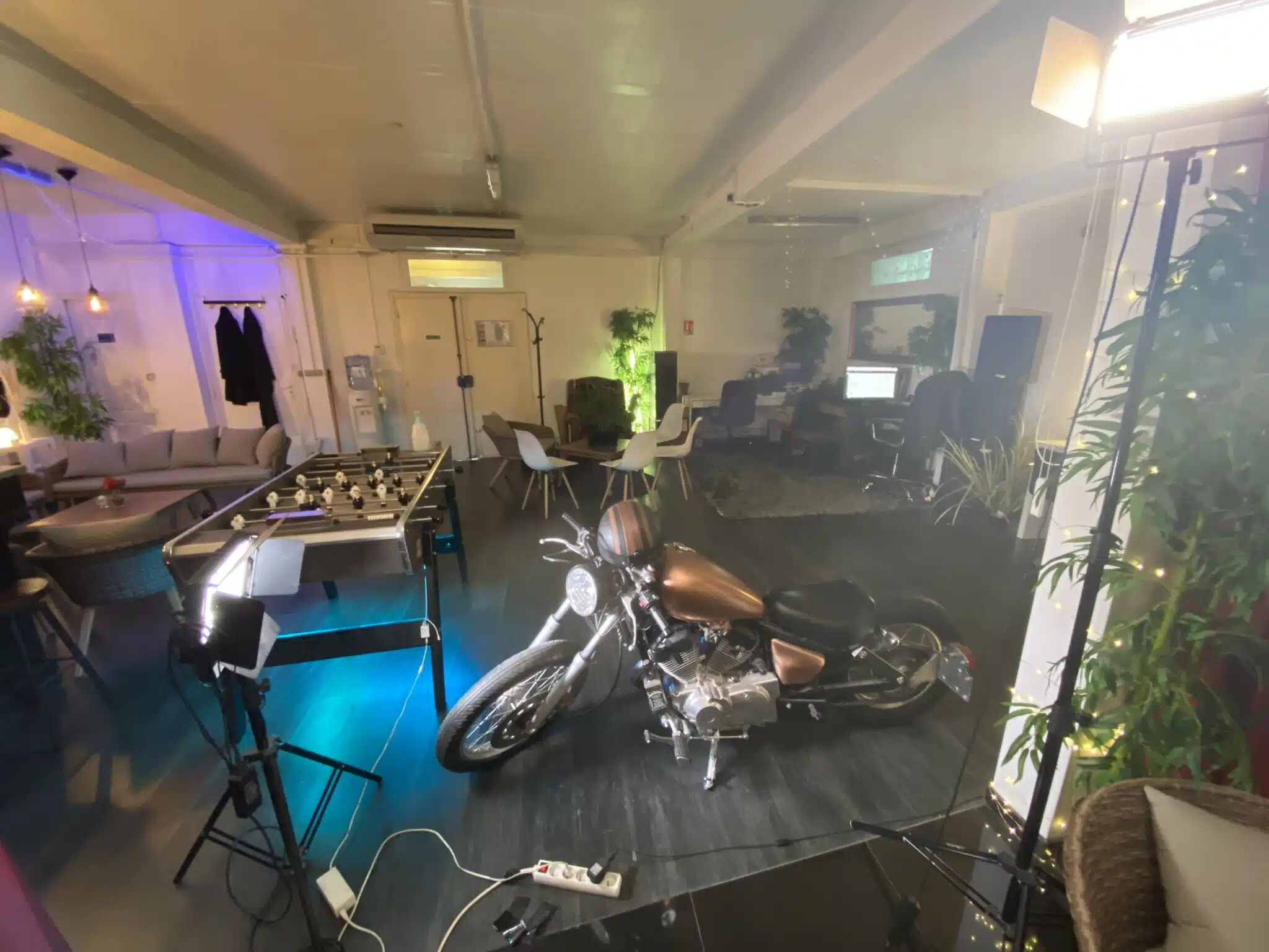 LE LOFT DU 20ÈME (75020) dispose d'un espace moderne et lumineux avec une moto centrale, un baby-foot, des canapés, des chaises de salle à manger, des bureaux d'ordinateur, des plantes en pot et du matériel d'éclairage de studio.