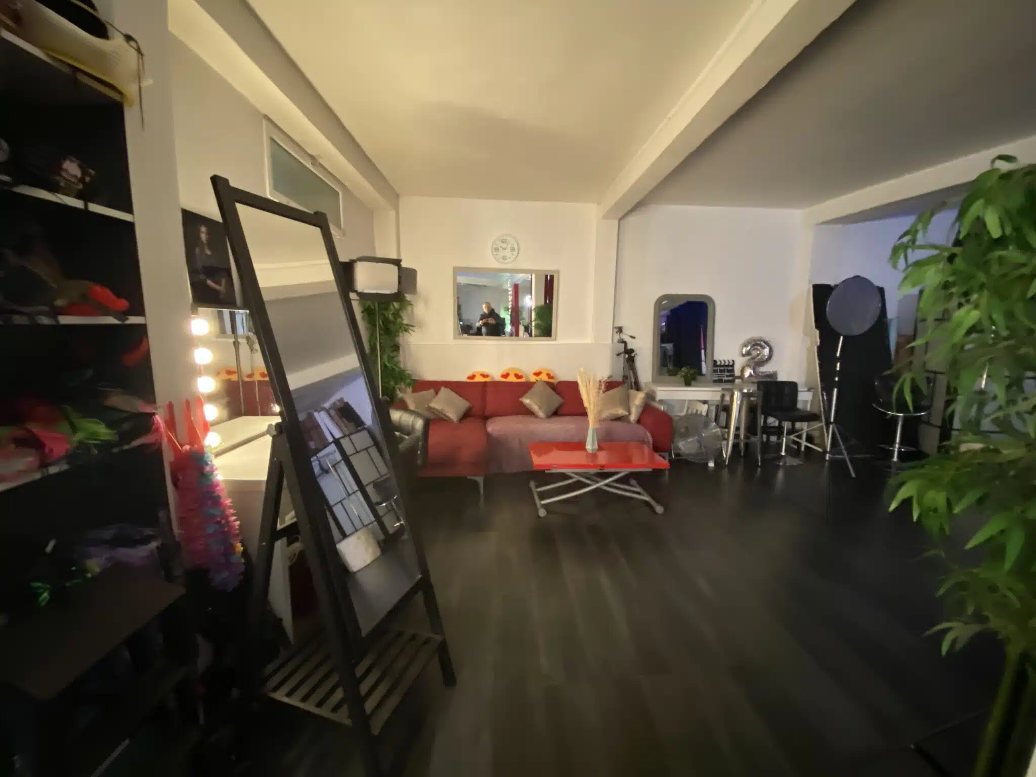 LE LOFT DU 20ÈME (75020) est un studio de photographie moderne avec un sol sombre, un canapé rouge, des miroirs avec des lampes de chevet, des accessoires, du matériel d'éclairage et une plante en pot sur le côté droit au premier plan.