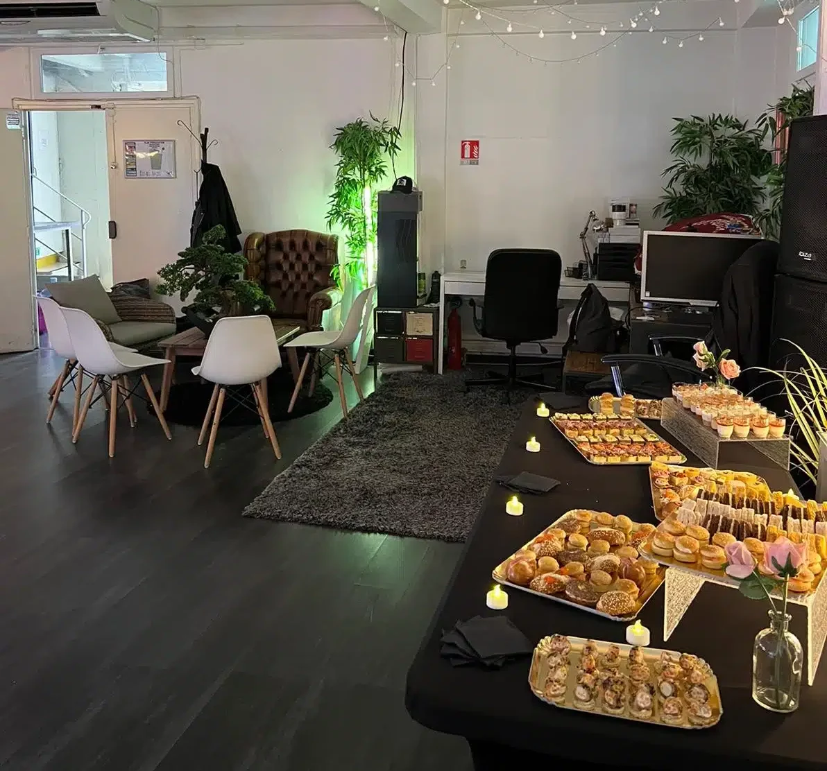 LE LOFT DU 20ÈME (75020) offre un espace intérieur confortable avec une table de bouchées assorties, des bougies, des chaises, des plantes et des meubles de bureau ; un éclairage doux et des guirlandes lumineuses créent une atmosphère accueillante.