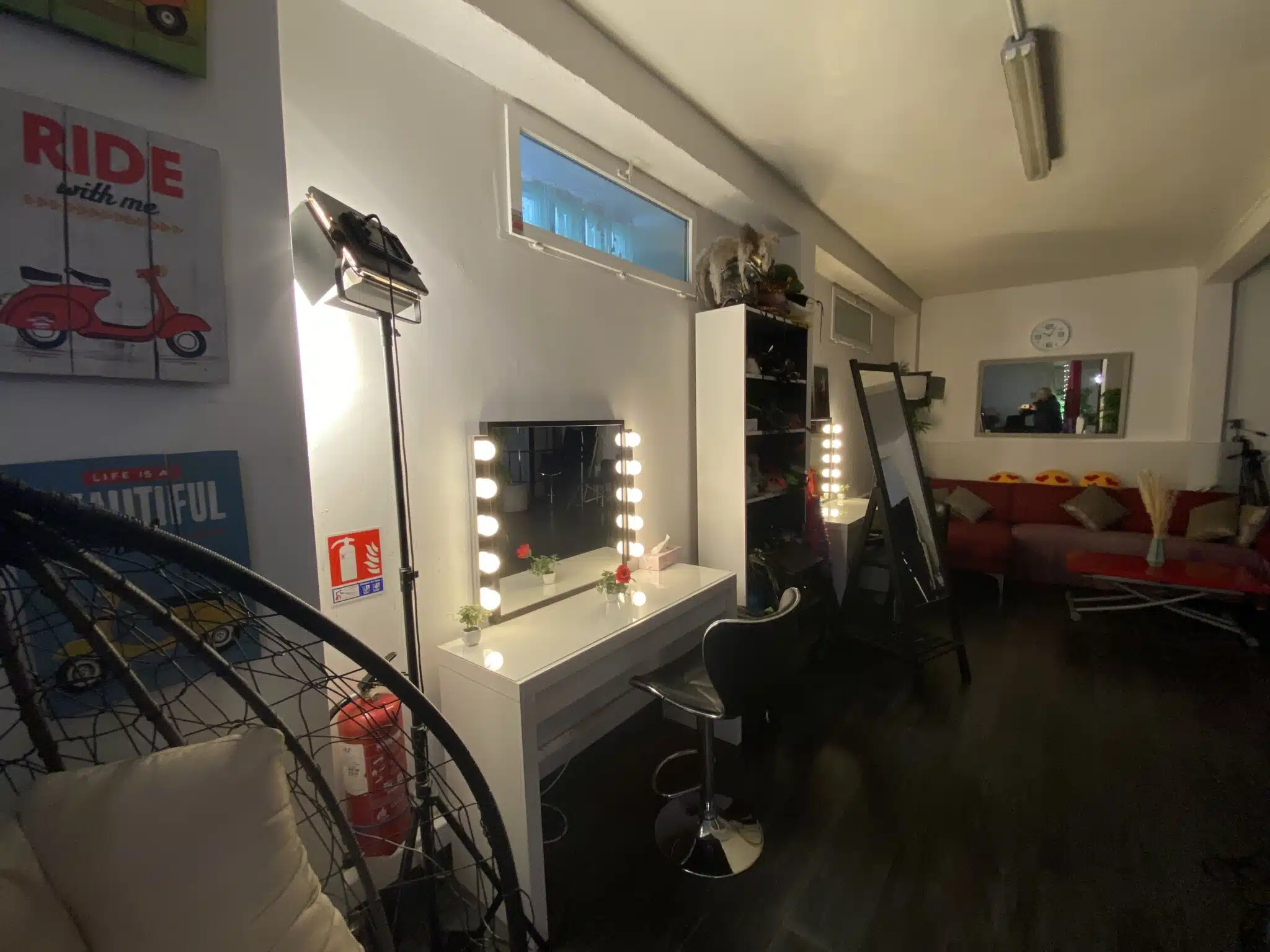 Une chambre confortable et moderne au LOFT DU 20ÈME (75020) comprend une station de maquillage bien éclairée avec un miroir, des lampes, une chaise noire, des affiches au mur, des étagères et un canapé rouge à l'arrière-plan.