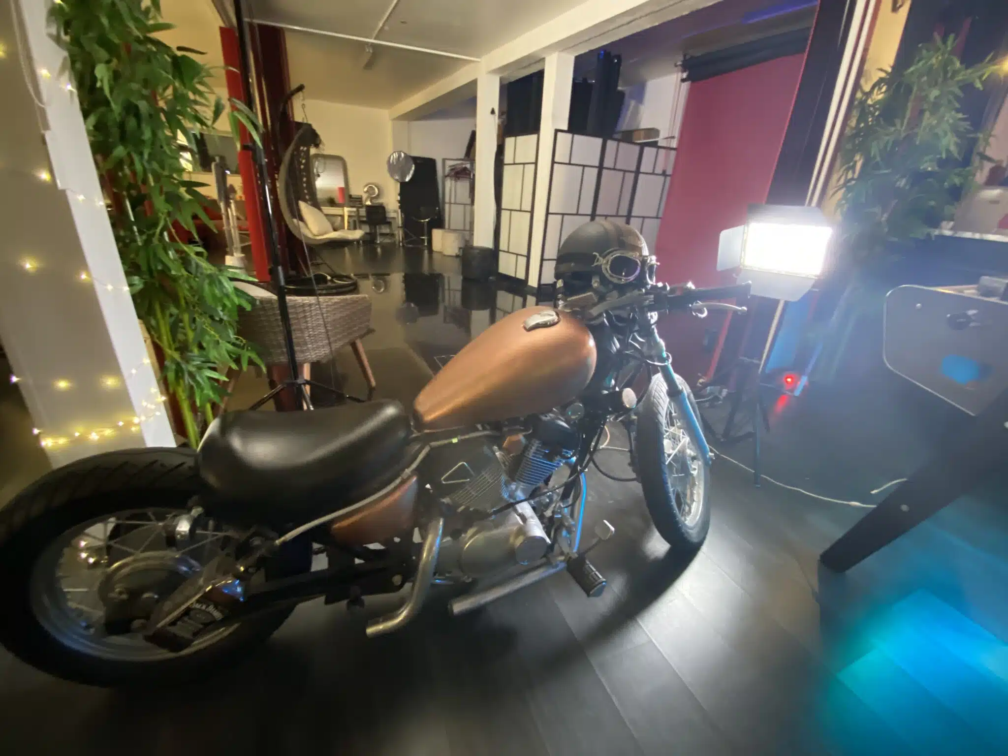 Une moto marron et noire avec un casque sur le guidon est garée à l'intérieur sur un sol sombre au LOFT DU 20ÈME (75020), entourée de plantes, de meubles et d'une lumière de studio.