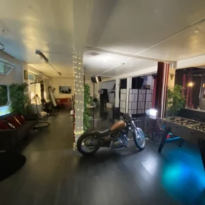 LE LOFT DU 20ÈME (75020) est un loft spacieux à la décoration moderne, avec une moto, un baby-foot, des canapés, des colonnes lumineuses, des plantes en pot et des lampes de studio pour une ambiance à la fois élégante et chaleureuse.