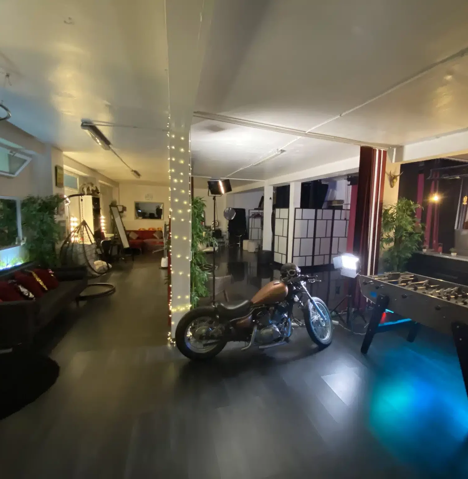 LE LOFT DU 20ÈME (75020) est un loft spacieux à la décoration moderne, avec une moto, un baby-foot, des canapés, des colonnes lumineuses, des plantes en pot et des lampes de studio pour une ambiance à la fois élégante et chaleureuse.