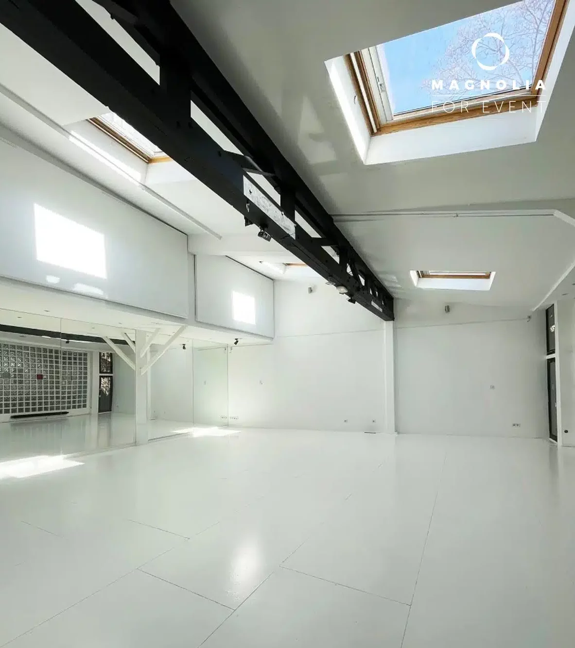 LE LOFT MONTMARTRE (75018) présente une salle blanche, spacieuse et moderne, avec de grandes fenêtres, des lucarnes, une poutre noire au plafond, une lumière naturelle abondante et un mur en miroir sur la gauche. Le logo Magnolia for Event apparaît dans le coin supérieur droit.