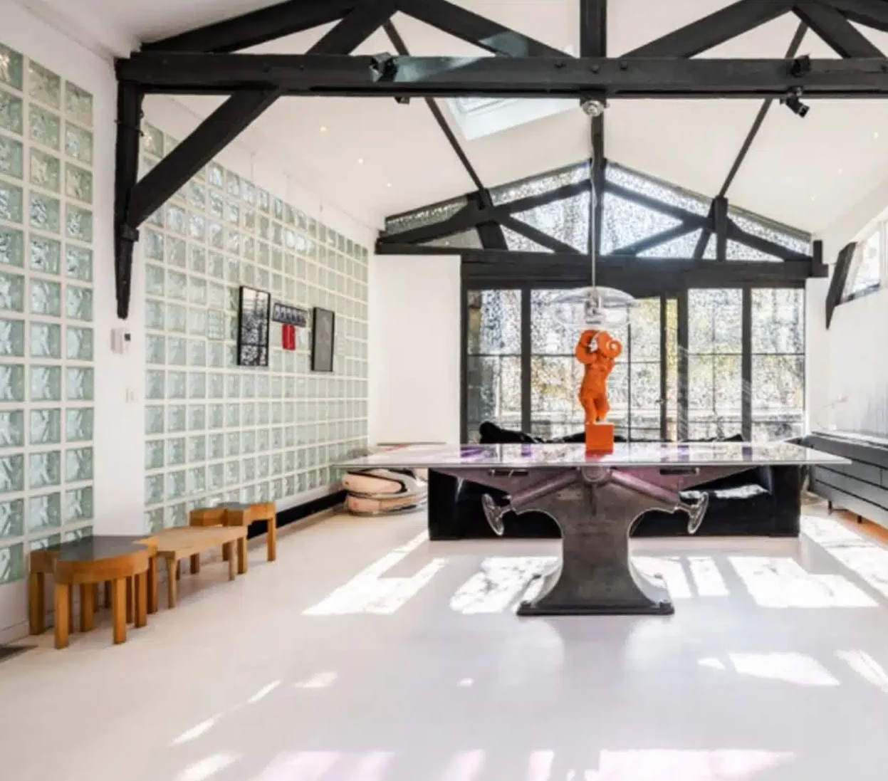 LE LOFT MONTMARTRE (75018) propose un espace ouvert moderne avec des poutres noires apparentes, un mur de briques en verre, une table centrale en verre avec une sculpture orange, des tabourets en bois, de grandes fenêtres et des sols blancs pour une ambiance lumineuse et aérée.