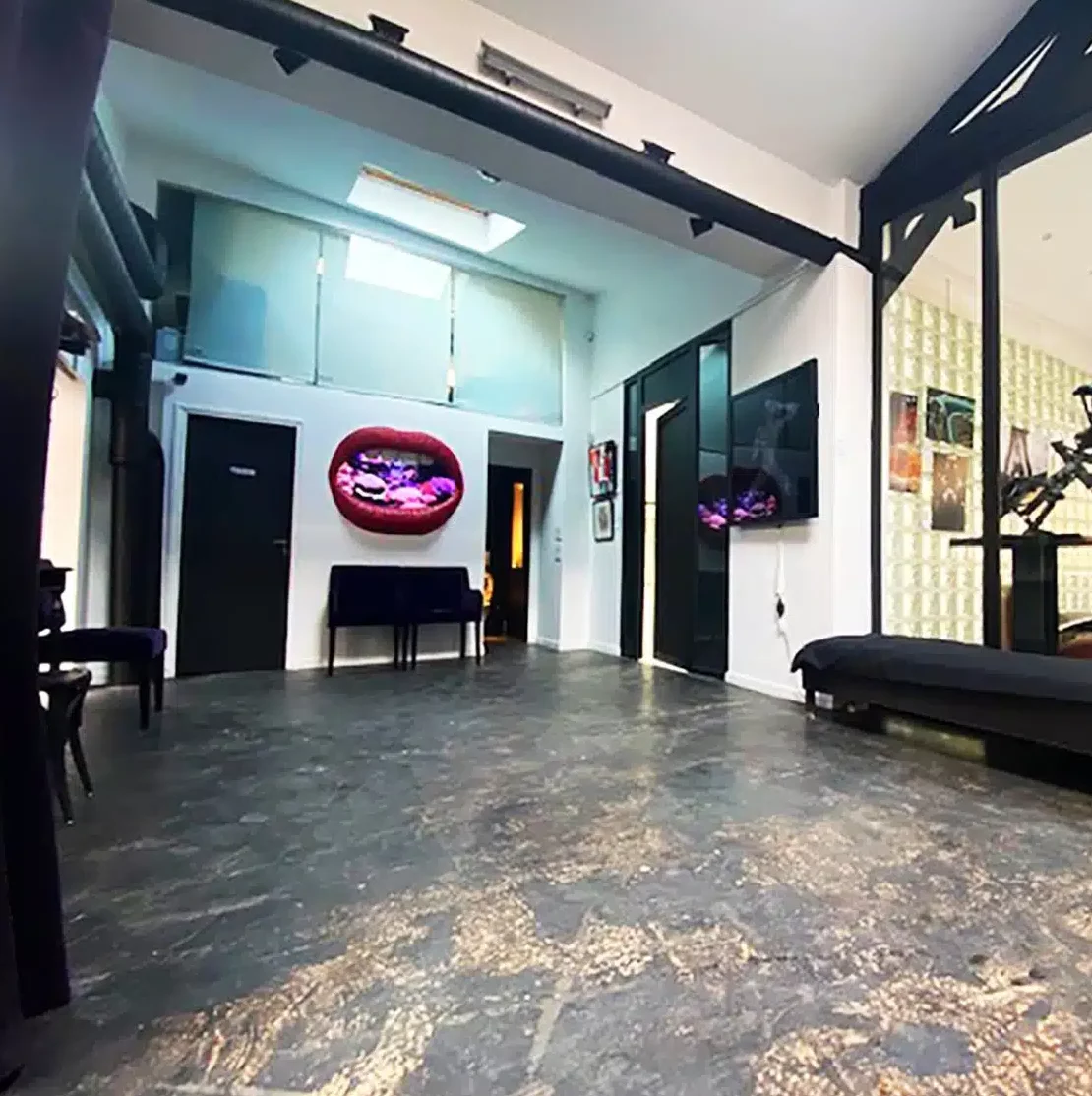 LE LOFT MONTMARTRE (75018) propose une pièce moderne avec des sols sombres, des murs blancs, des poutres noires, une télévision à écran plat, une œuvre d'art illuminée, une banquette noire, des cloisons en verre, un puits de lumière au-dessus, et une atmosphère propre et ouverte.