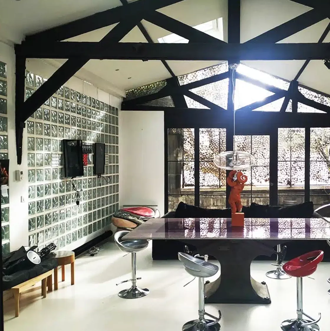 LE LOFT MONTMARTRE (75018) dispose d'une salle à manger moderne avec des poutres noires apparentes au plafond, un mur de blocs de verre, une table élégante, des tabourets de bar dynamiques et de grandes fenêtres. Une sculpture décorative orange agrémente l'espace.