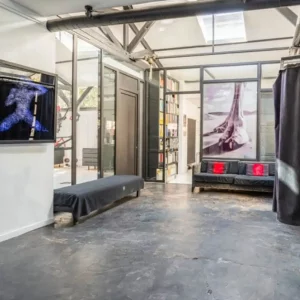LE LOFT MONTMARTRE (75018) : Pièce industrielle moderne avec sols en béton gris, murs en verre, canapé noir avec coussins rouges, banc, art mural, tuyaux apparents, et lumière naturelle provenant de grandes fenêtres et de lucarnes.