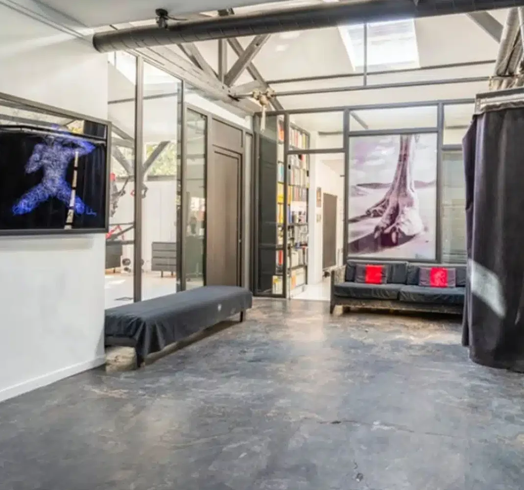 LE LOFT MONTMARTRE (75018) : Pièce industrielle moderne avec sols en béton gris, murs en verre, canapé noir avec coussins rouges, banc, art mural, tuyaux apparents, et lumière naturelle provenant de grandes fenêtres et de lucarnes.
