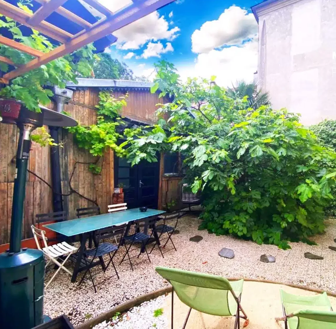 LE LOFT MONTMARTRE (75018) dispose d'un patio extérieur confortable avec une table verte, des chaises noires et blanches, deux chaises longues vertes, un sol en gravier, de la verdure luxuriante et un bâtiment en bois sous un ciel bleu partiellement nuageux.