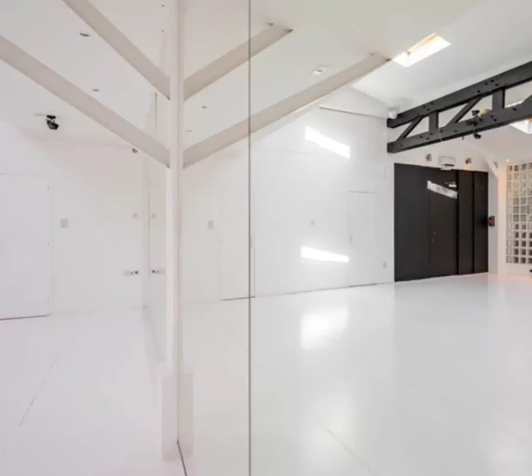 LE LOFT MONTMARTRE (75018) présente un intérieur spacieux et moderne avec des sols, des murs et des plafonds blancs, séparés par un mur en miroir. Les poutres noires et les blocs de verre offrent un contraste saisissant avec l'espace minimaliste et lumineux.