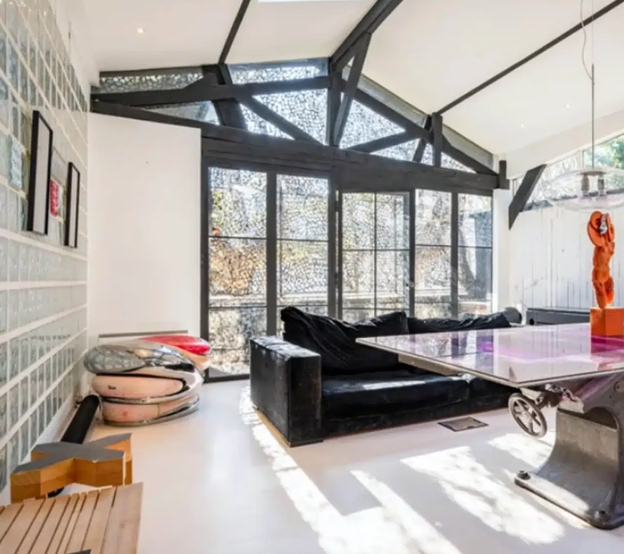 LE LOFT MONTMARTRE (75018) dispose d'un salon moderne avec de grandes fenêtres, un canapé noir, un mur en briques de verre, une décoration unique, une table en verre avec une sculpture orange, et une lumière naturelle brillante.
