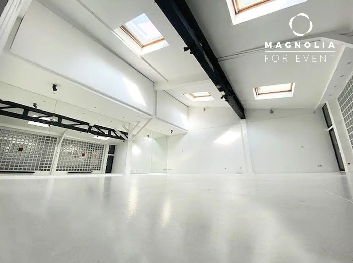Salle blanche spacieuse et moderne au LOFT MONTMARTRE (75018) avec de hauts plafonds, des puits de lumière, des poutres noires et de grands blocs de verre. La salle vide est éclairée par la lumière naturelle et présente un décor minimaliste. Le logo de Magnolia For Event se trouve dans le coin supérieur droit.