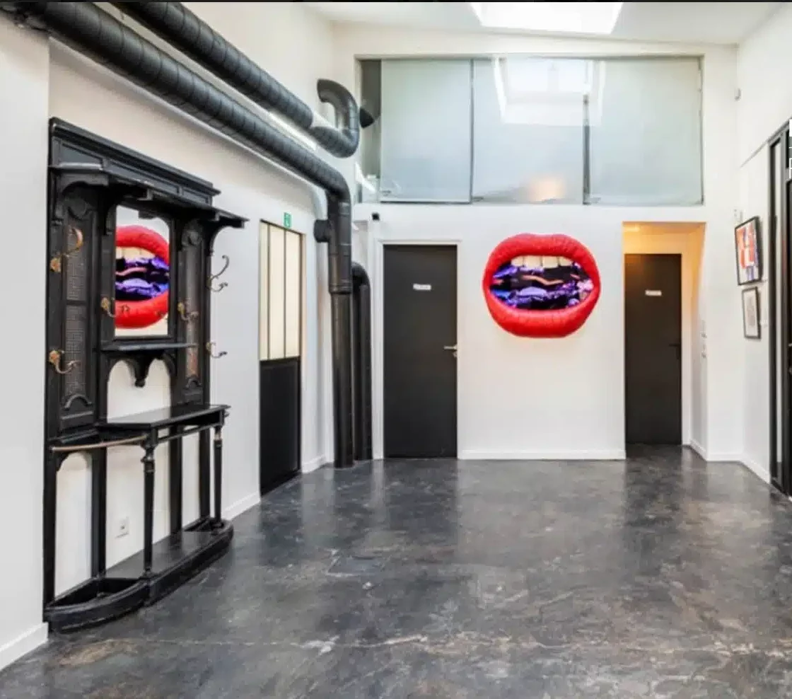 LE LOFT MONTMARTRE (75018) présente un couloir moderne avec des sols sombres, des murs blancs, des portes noires fermées, des lèvres rouges, un miroir noir décoratif reflétant les œuvres d'art sur la gauche, et des tuyaux de plafond apparents.