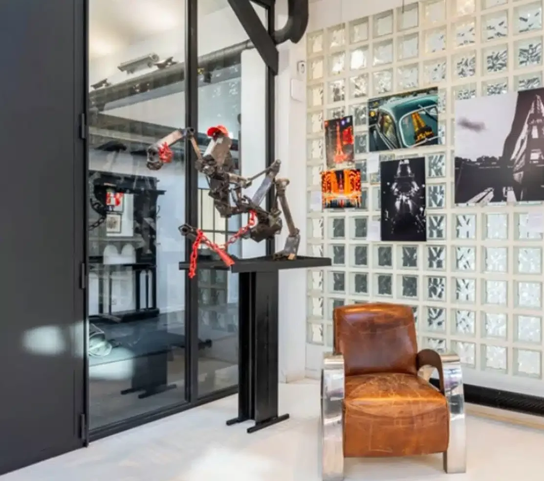 Une chambre moderne à LE LOFT MONTMARTRE (75018) comprend une chaise en cuir brun et en métal, un mur en verre, une sculpture abstraite en métal sur un piédestal, et des murs ornés de photographies et de blocs de verre.