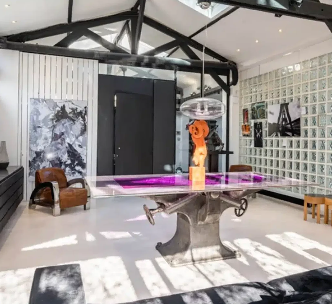 LE LOFT MONTMARTRE (75018) présente une ambiance industrielle moderne avec des poutres noires apparentes, des fenêtres en verre, des fauteuils en cuir, de l'art abstrait et une table sculpturale avec un centre de table orange. Des puits de lumière illuminent l'espace.
