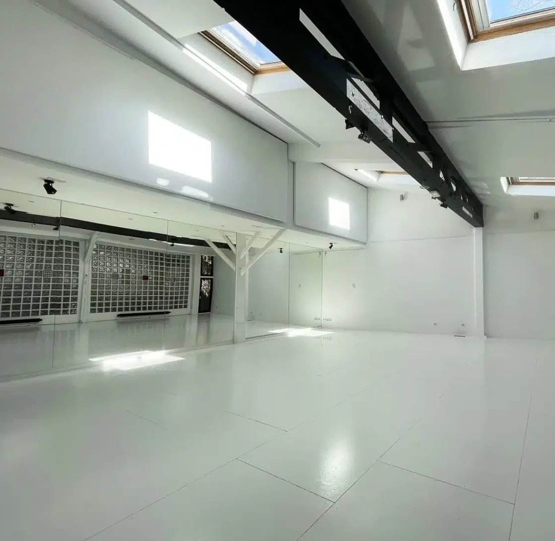 LE LOFT MONTMARTRE (75018) est un studio spacieux et moderne avec des sols et des murs blancs, de grandes fenêtres et des lucarnes pour la lumière naturelle, des poutres apparentes et un mur en miroir sur un côté.